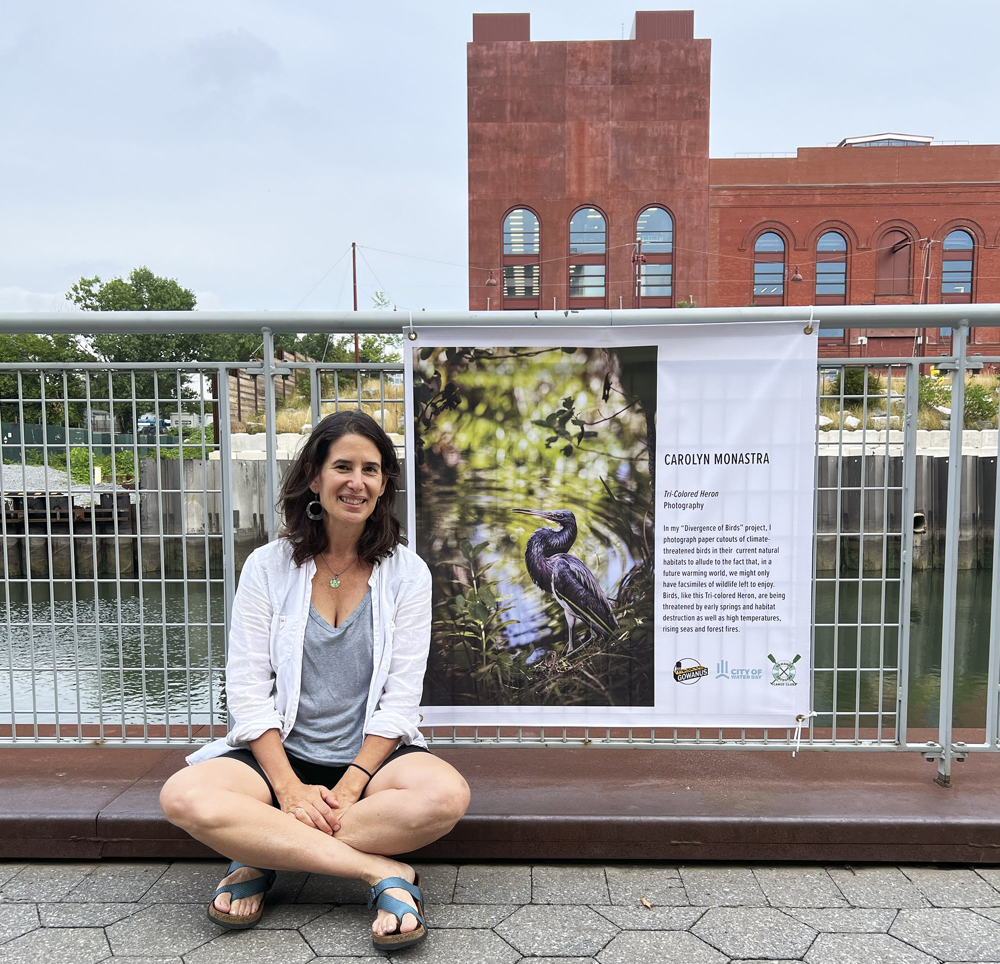 The artist sits in front of a banner with her image of a “Tri-colored Heron.” This was displayed along the Gowanus Canal as part of the City of Water Day in New York City, Summer 2022.